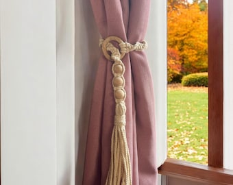 May include: A beige macrame curtain tieback with wooden beads and a tassel. The tieback is holding back a dusty rose curtain.