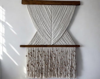 Könnte beinhalten: Ein weißer Makramee-Wandbehang mit geometrischem Muster und Holzleisten. Der untere Teil des Behangs ist mit einer Fransenborte mit Holzperlen verziert.