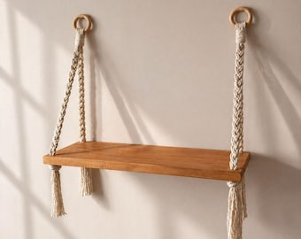 May include: A wooden floating shelf with a natural finish, suspended by braided cream-colored rope. The shelf is rectangular, and the rope is attached to wooden rings at the top. Tassels hang from the bottom of the shelf. The shelf is designed for wall mounting.