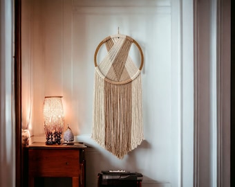 May include: A macrame wall hanging with a wooden ring and beige and white yarn. The hanging is on a white wall with a lamp on a wooden table in the foreground.