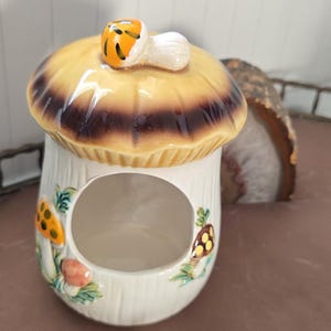 May include: A ceramic mushroom-shaped container with a yellow and brown cap and a white stem. The container has a large opening and is decorated with colorful mushroom and leaf designs. A small mushroom adorns the top.