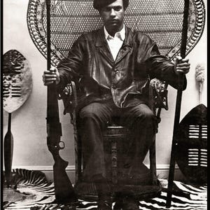 May include: Black and white photo of Huey P. Newton, a prominent figure in the Black Panther Party, seated on a throne-like chair with a rifle and spear. He is wearing a leather jacket and a beret.