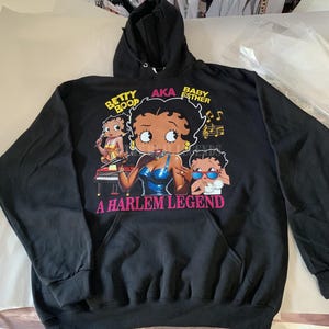 May include: Black hooded sweatshirt with a graphic print of Betty Boop playing the piano with the text "Betty Boop AKA Baby Esther A Harlem Legend".