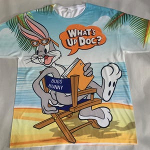 May include: White t-shirt with a colorful print of Bugs Bunny sitting in a director's chair on a beach. The text "What's Up Doc?" is in a speech bubble above Bugs Bunny. The text "Bugs Bunny" is on the back of the director's chair.