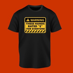 May include: Black t-shirt with a yellow warning sign graphic. The sign reads "WARNING THESE HANDS RATED 'E' -FOR EVERYONE". The shirt has a crew neck and short sleeves. The background is a solid orange color.