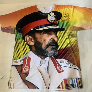 May include: A white t-shirt with a colorful graphic of a man in a military uniform and hat. The graphic has a yellow, orange, and green background. The text "Haile Selassie I" is printed on the shirt.