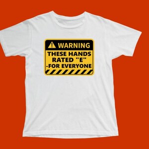 May include: White t-shirt with a yellow and black warning sign that reads "WARNING THESE HANDS RATED 'E' -FOR EVERYONE". The shirt has a crew neck and short sleeves. The background is a solid orange color.