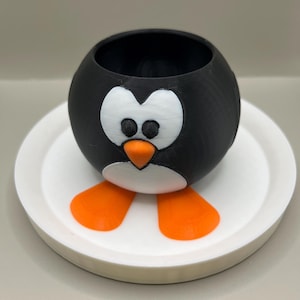 May include: A black and white penguin-shaped planter with orange feet and beak, sitting on a white plate. The planter has a round body with a large opening at the top for planting. The design is whimsical and fun.