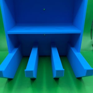 May include: A bright blue wall-mounted storage organizer featuring a rectangular top shelf and four protruding vertical dividers extending from the base. The unit has two small circular mounting holes centered on the back panel for installation.