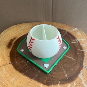 May include: A white baseball-themed container with red stitching details, divided into two compartments. It sits on a baseball field-shaped base with a brown infield, green outfield, and white home plate and bases. The container is on a wooden surface.