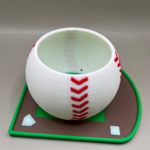 May include: White ceramic baseball planter with red stitching, sitting on a green and brown baseball field base.