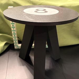 May include: A black side table with a round top featuring a white circle with the number 8. The table has three black legs and is likely made of wood or a similar material.