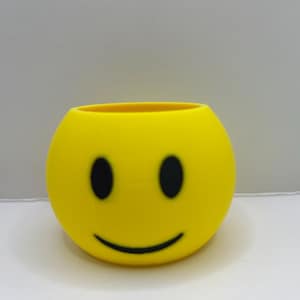 May include: A yellow smiley face planter with black eyes and a curved mouth. The round planter has a smooth surface and a hollow interior. The planter is designed to hold small plants or other decorative items.