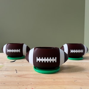 May include: Three brown and white football-shaped planters with green bases. The planters are arranged in a row on a wooden surface.
