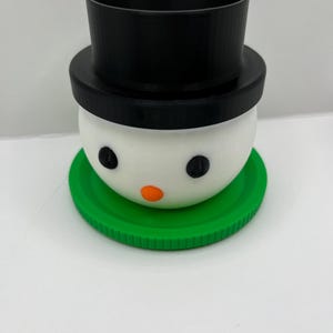 May include: A snowman-shaped container with a black top hat, white face, and green base. The snowman has black eyes and an orange nose. The container is designed to hold small items or serve as a decorative piece.