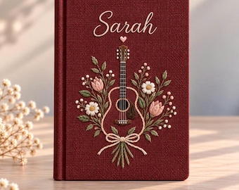 May include: A deep red fabric-covered journal featuring an embroidered acoustic guitar surrounded by a bouquet of white and pink flowers with green leaves. The name Sarah is stitched in white script above the guitar with a small heart.