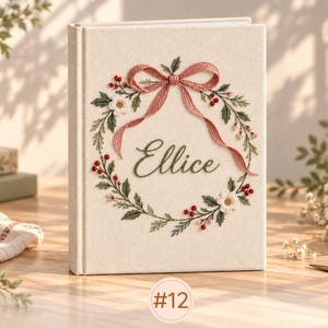 May include: A cream-colored fabric-covered book featuring an embroidered wreath design with green leaves, small red berries, and white flowers. A pink embroidered bow sits at the top of the wreath, and the name Ellice is stitched in dark green script.