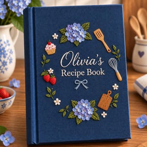 May include: A navy blue hardback recipe book with the text Olivia's Recipe Book in white. The cover features embroidered designs of blue hydrangea flowers, green leaves, a cupcake, strawberries, a wooden spatula, a whisk, and a cutting board.