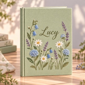 May include: A sage green fabric-covered book featuring an embroidered floral arrangement of white daisies, blue hydrangeas, purple lavender, and blue bellflowers. The name Lucy is embroidered in a dark green cursive script in the centre.