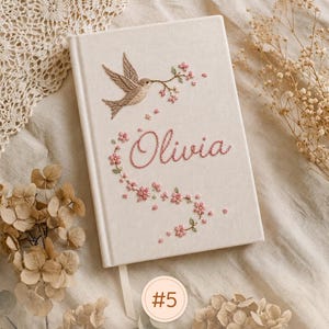 Pode incluir: Um di&aacute;rio de capa dura na cor creme com um design bordado de um p&aacute;ssaro segurando um ramo florido. O nome Olivia est&aacute; bordado em linha rosa abaixo do p&aacute;ssaro, rodeado por pequenos detalhes florais cor-de-rosa e uma fita marcadora.