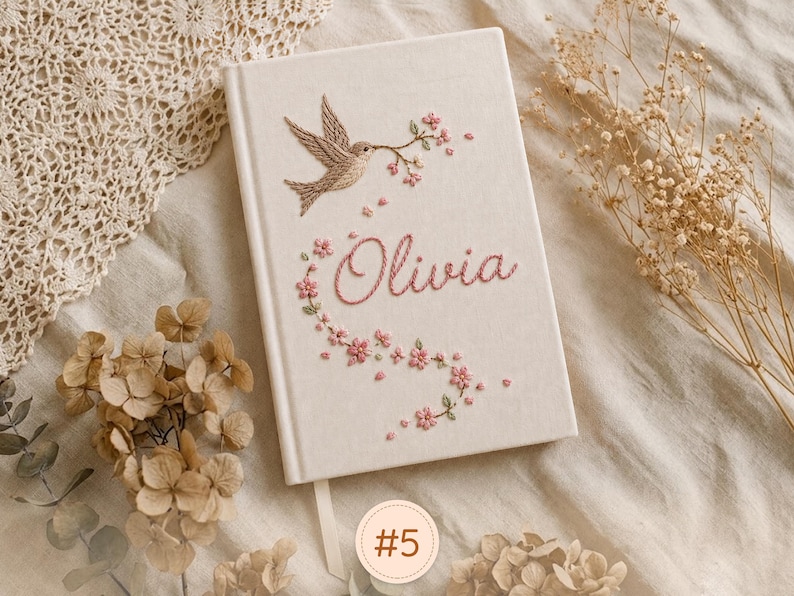 Pode incluir: Um di&aacute;rio de capa dura na cor creme com um design bordado de um p&aacute;ssaro segurando um ramo florido. O nome Olivia est&aacute; bordado em linha rosa abaixo do p&aacute;ssaro, rodeado por pequenos detalhes florais cor-de-rosa e uma fita marcadora.