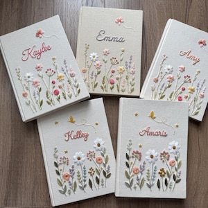 May include: Five off-white fabric-covered journals featuring embroidered wildflower designs and personalized names in cursive script. Each book displays unique floral patterns in pink, yellow, and green threads, with small embroidered butterflies or stars.