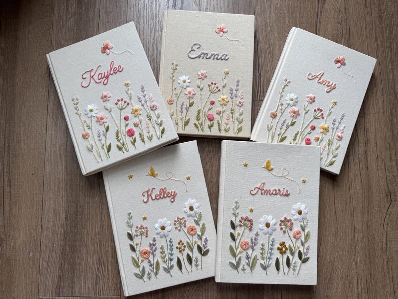 May include: Five off-white fabric-covered journals featuring embroidered wildflower designs and personalized names in cursive script. Each book displays unique floral patterns in pink, yellow, and green threads, with small embroidered butterflies or stars.
