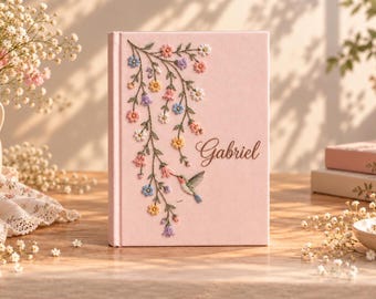 May include: A light pink hardcover book featuring an embroidered design of trailing vines with colorful flowers and a small hummingbird. The name Gabriel is embroidered in a dark script font on the right side of the front cover.