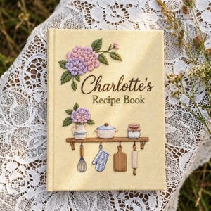 May include: A cream-coloured hardback recipe book featuring embroidered details of pink and purple hydrangea flowers, a blue and white pot, a jar, a whisk, a blue chequered oven mitt, a wooden cutting board, and a rolling pin with the text Charlotte's Recipe Book.