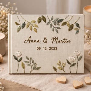 May include: A cream-coloured fabric-covered photo album featuring embroidered green leaf motifs and white flowers. The cover displays the text Anna & Martin and the date 09.12.2023 in a dark brown script font, centred on the front panel.