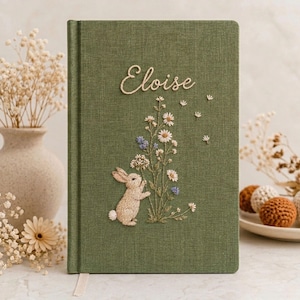 May include: A sage green fabric-covered journal featuring an embroidered design of a small rabbit standing near a cluster of wildflowers. The name Eloise is embroidered in white script above the floral arrangement on the front cover.