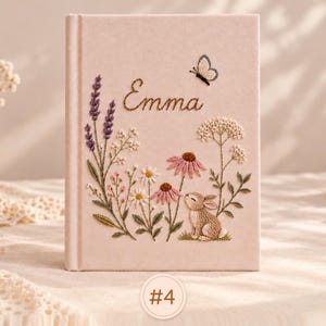 May include: A light pink hardcover journal featuring intricate embroidery of lavender, daisies, coneflowers, a small brown rabbit, and a butterfly. The name Emma is embroidered in brown script above the floral design. A small circular tag labeled #4 is visible.