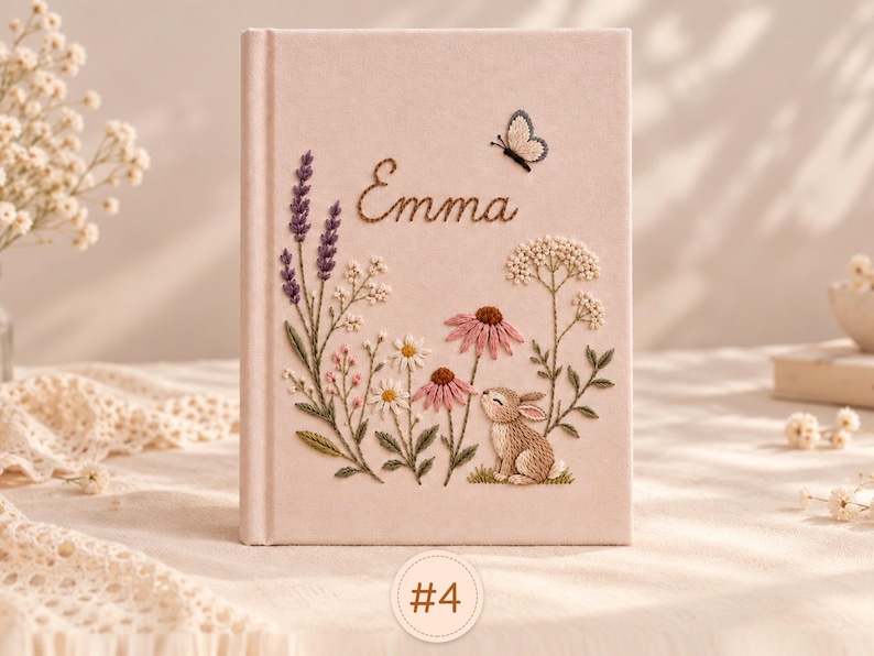 May include: A light pink hardcover journal featuring intricate embroidery of lavender, daisies, coneflowers, a small brown rabbit, and a butterfly. The name Emma is embroidered in brown script above the floral design. A small circular tag labeled #4 is visible.