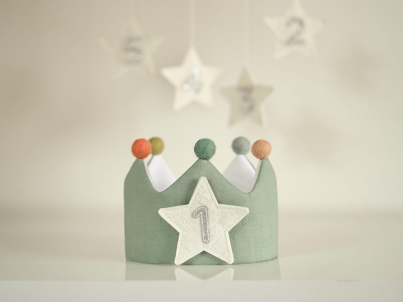 Op de afbeelding: Een groene viltkroon met een witte ster en het cijfer "1" erop. De kroon heeft vijf gekleurde viltballetjes bovenop.