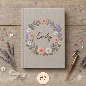 Pode incluir: Um di&aacute;rio revestido de tecido bege claro com uma coroa de flores bordada em tons de rosa, branco e roxo, envolvendo o nome Emily em letra cursiva castanha. O di&aacute;rio &eacute; acompanhado por ramos de lavanda seca e uma caneta de caligrafia.