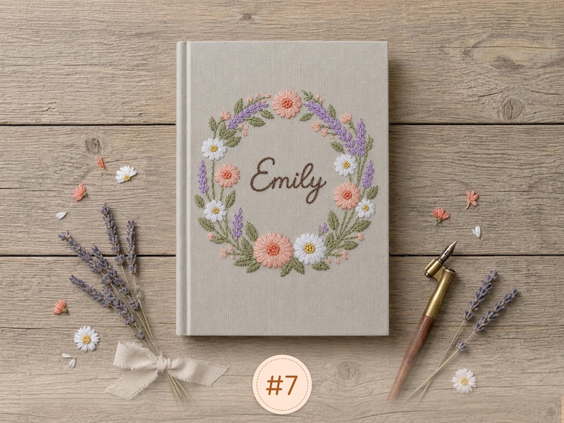 Pode incluir: Um di&aacute;rio revestido de tecido bege claro com uma coroa de flores bordada em tons de rosa, branco e roxo, envolvendo o nome Emily em letra cursiva castanha. O di&aacute;rio &eacute; acompanhado por ramos de lavanda seca e uma caneta de caligrafia.