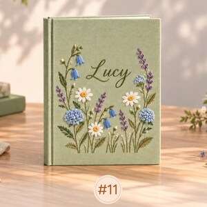 May include: A sage green hardcover book featuring embroidered floral designs in shades of blue, purple, and white. The name Lucy is stitched in dark green cursive script in the center. The cover displays various wildflowers and stems.