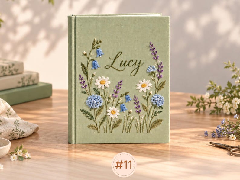 May include: A sage green hardcover book featuring embroidered floral designs in shades of blue, purple, and white. The name Lucy is stitched in dark green cursive script in the center. The cover displays various wildflowers and stems.