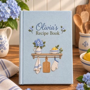 Könnte beinhalten: Ein hellblaues Hardcover-Buch mit dem Titel Olivia's Recipe Book und gestickten Details auf dem Einband. Das Motiv zeigt ein Holzregal mit einem Krug voller blauer Blumen, einer Schale mit Zitronen, einem Tuch, einem Schneidebrett und einem Ofenhandschuh.