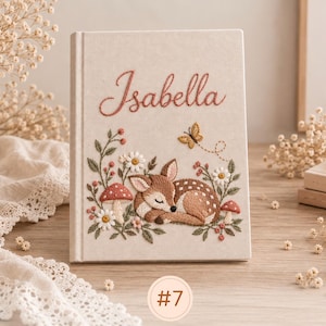 May include: A cream-colored notebook featuring an embroidered design of a sleeping fawn surrounded by mushrooms, white flowers, and a small butterfly. The name Isabella is embroidered in a cursive script at the top of the cover.