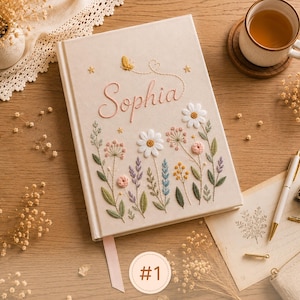 Pode incluir: Um di&aacute;rio de capa dura na cor creme com desenhos florais bordados em tons de branco, rosa, azul e verde. O nome Sophia est&aacute; bordado com linha rosa acima das flores, acompanhado por uma pequena borboleta amarela e estrelas.