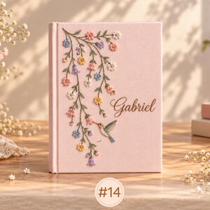 May include: A light pink hardcover book featuring an embroidered design of trailing flowers in shades of blue, yellow, and pink, a small hummingbird, and the name Gabriel written in elegant brown cursive script on the front cover.