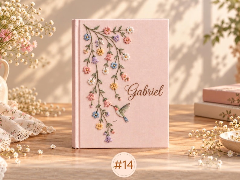 May include: A light pink hardcover book featuring an embroidered design of trailing flowers in shades of blue, yellow, and pink, a small hummingbird, and the name Gabriel written in elegant brown cursive script on the front cover.