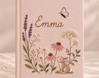 May include: A light pink fabric-covered journal featuring hand-embroidered floral designs, including purple lavender, white daisies, and pink coneflowers. A small brown rabbit sits among the flowers, and a butterfly is embroidered above the name Emma.