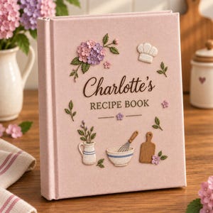 May include: A light pink fabric-covered recipe book titled Charlotte's RECIPE BOOK. The cover features embroidered designs of purple hydrangea flowers, a chef's hat, a small pitcher, a mixing bowl with a whisk, and a wooden cutting board.