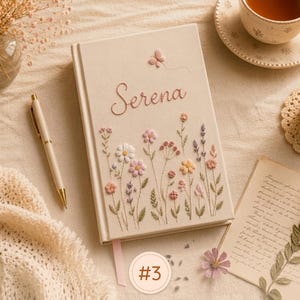 Pode incluir: Um di&aacute;rio de capa dura na cor creme com flores silvestres bordadas em tons de rosa, amarelo e roxo, com o nome Serena costurado em letra cursiva e uma pequena borboleta acima. Uma caneta creme a condizer repousa ao lado do di&aacute;rio.