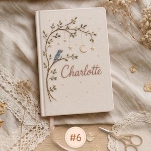 Pode incluir: Um di&aacute;rio de capa dura na cor creme com um design bordado de um p&aacute;ssaro azul pousado num ramo com folhas, estrelas pendentes e uma lua crescente. O nome Charlotte est&aacute; bordado em letras castanhas na capa frontal.