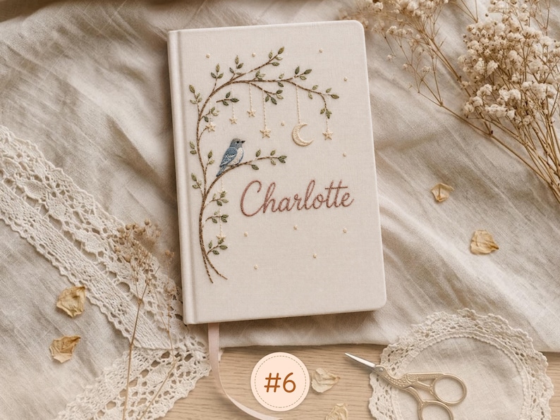 Pode incluir: Um di&aacute;rio de capa dura na cor creme com um design bordado de um p&aacute;ssaro azul pousado num ramo com folhas, estrelas pendentes e uma lua crescente. O nome Charlotte est&aacute; bordado em letras castanhas na capa frontal.