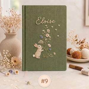 Pode incluir: Um di&aacute;rio retangular revestido de tecido verde-oliva com um desenho bordado de um pequeno coelho em p&eacute; ao lado de um grupo de flores silvestres brancas e azuis. O nome Eloise est&aacute; bordado em letra cursiva branca no topo da capa.