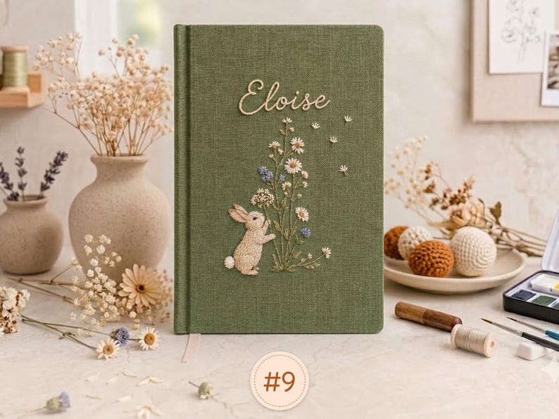 Pode incluir: Um di&aacute;rio retangular revestido de tecido verde-oliva com um desenho bordado de um pequeno coelho em p&eacute; ao lado de um grupo de flores silvestres brancas e azuis. O nome Eloise est&aacute; bordado em letra cursiva branca no topo da capa.